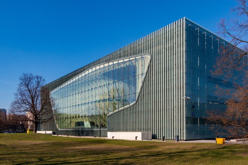 Modern building with large glass fronts and vertical structures on the facade, surrounded by lawns and trees, in sunny weather.
