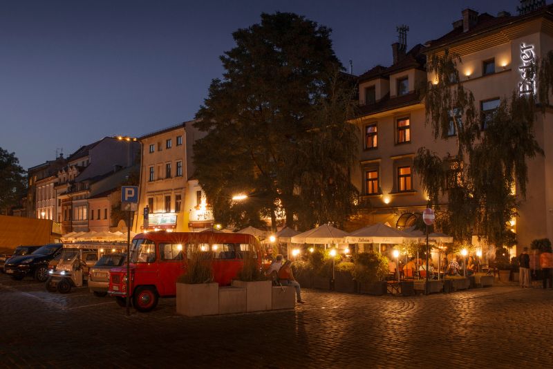 Wieczorna scena z Krakowa, przedstawiająca oświetlony plac z restauracjami i zaparkowanym czerwonym busem, otoczonym parasolami i ludźmi siedzącymi na zewnątrz.