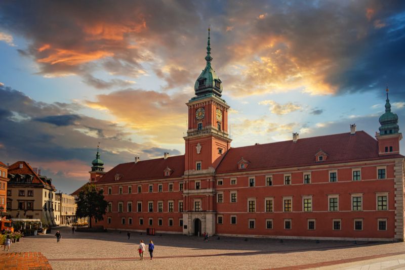 The Royal Castle in Warsaw at sunset, with baroque facade and clock tower, against dramatic clouds in the sky.