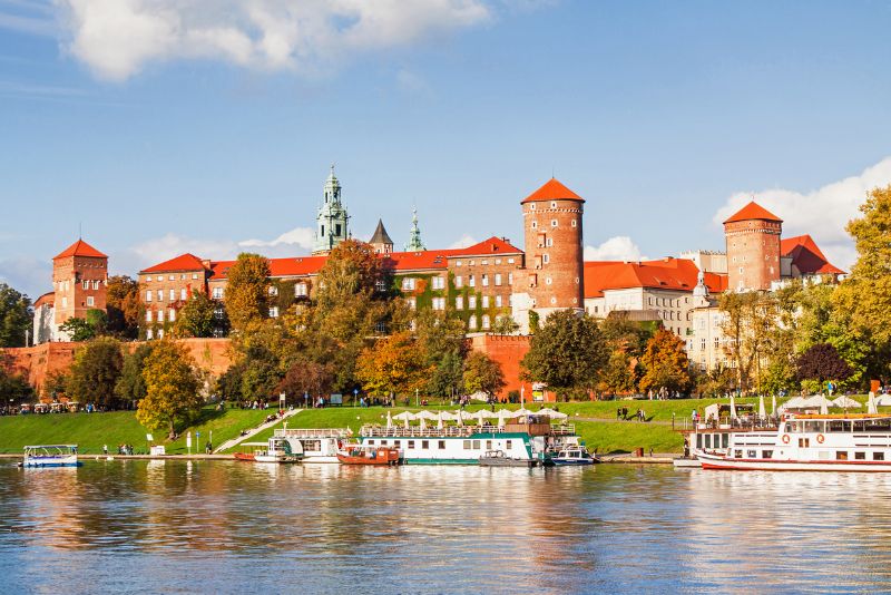 Panorama Wawelu w Krakowie, z widokiem na ceglany zamek z czerwonymi dachami, otoczony zielenią; przed zamkiem płynie rzeka Wisła z kilkoma łodziami zacumowanymi przy brzegu.