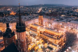 Nocny widok na krakowski Rynek Główny z oświetlonym Sukiennicami i tłumem ludzi na placu, w tle zarysowane miasto i oświetlone ulice.