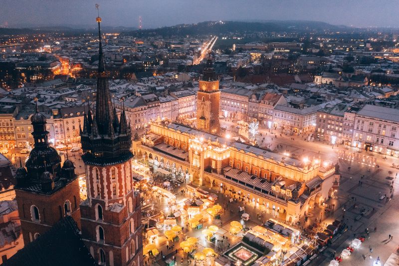 Nocny widok na krakowski Rynek Główny z oświetlonym Sukiennicami i tłumem ludzi na placu, w tle zarysowane miasto i oświetlone ulice.