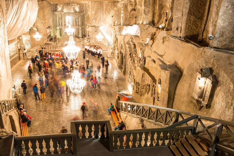 Podziemna kaplica w Kopalni Soli w Wieliczce, oświetlona dużymi żyrandolami, z widocznymi rzeźbami na ścianach, gośćmi i chórem stojącym na podwyższeniu.