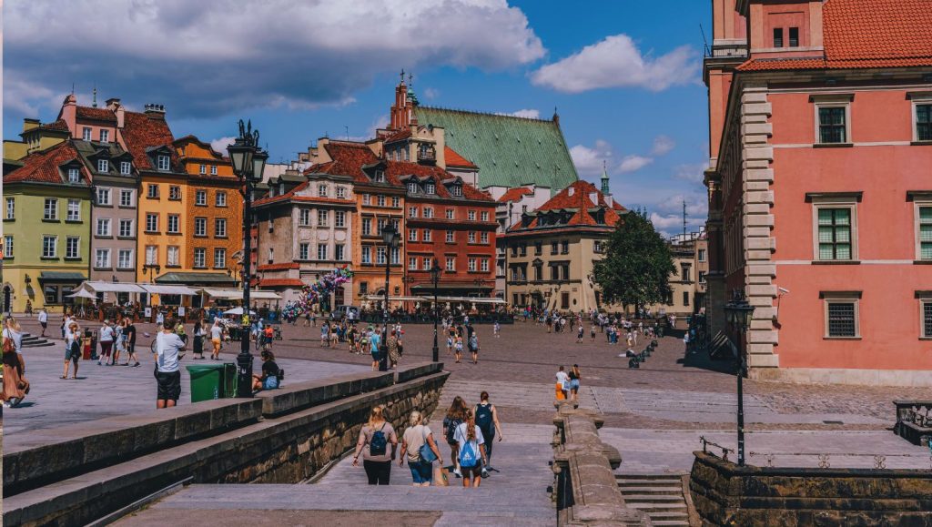 Plac Zamkowy w Warszawie z kolorowymi kamienicami i ludźmi spacerującymi po bruku w pogodny dzień.