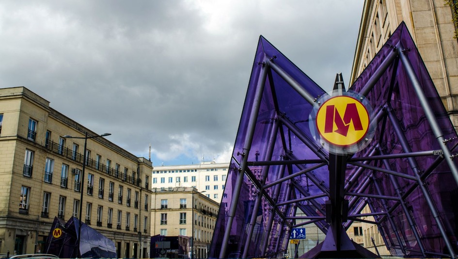 An entrance to a metro station with a purple structure and metro symbol, surrounded by urban architecture against a cloudy sky.