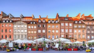 A view of townhouses in the Old Town in Warsaw, with outdoor restaurants under white umbrellas, surrounded by flowers, on a sunny day.