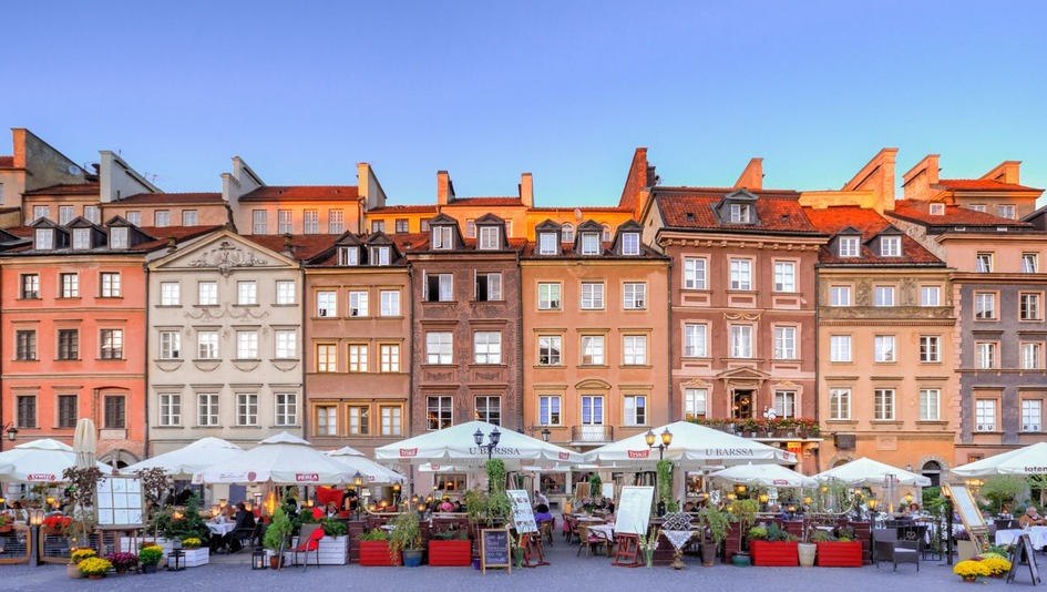 A view of townhouses in the Old Town in Warsaw, with outdoor restaurants under white umbrellas, surrounded by flowers, on a sunny day.