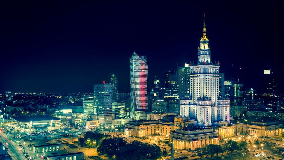 A view of the Palace of Culture and Science in Warsaw illuminated at night and the modern skyscrapers surrounding it.