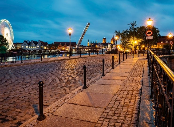 A night view of a cobblestone street with illuminated lanterns, against the backdrop of a drawbridge and a Ferris wheel in the background.