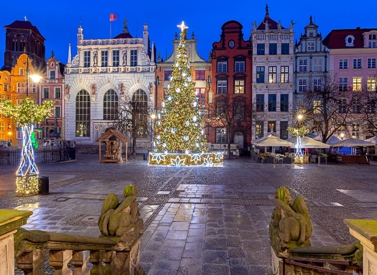 An illuminated Christmas tree in a square in Gdańsk, surrounded by historic, colorful buildings in the background, visible against the night sky.