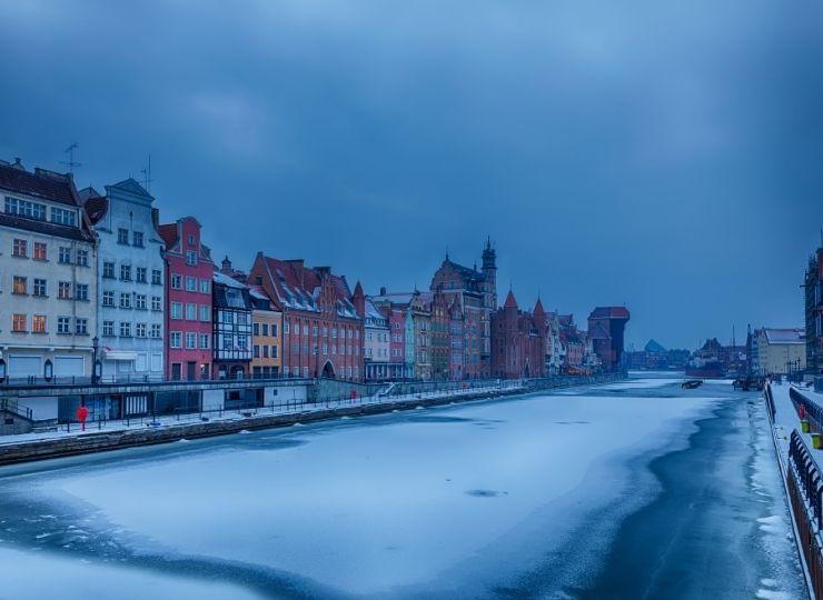 A winter view of the frozen Motława River in Gdańsk, with a row of colorful, historic townhouses on the left side, under a cloudy sky.