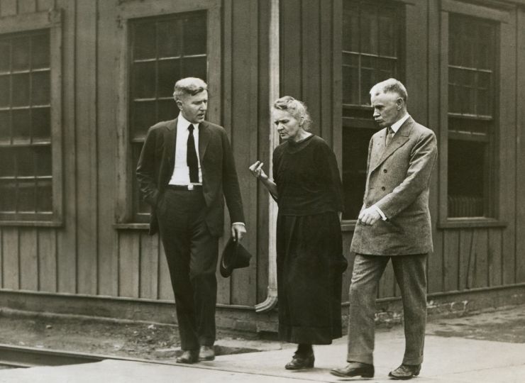 Three people walking on a sidewalk in front of a wooden building. The central figure, Marie Curie, is wearing a long, dark dress, and on either side of her are two men in suits.
