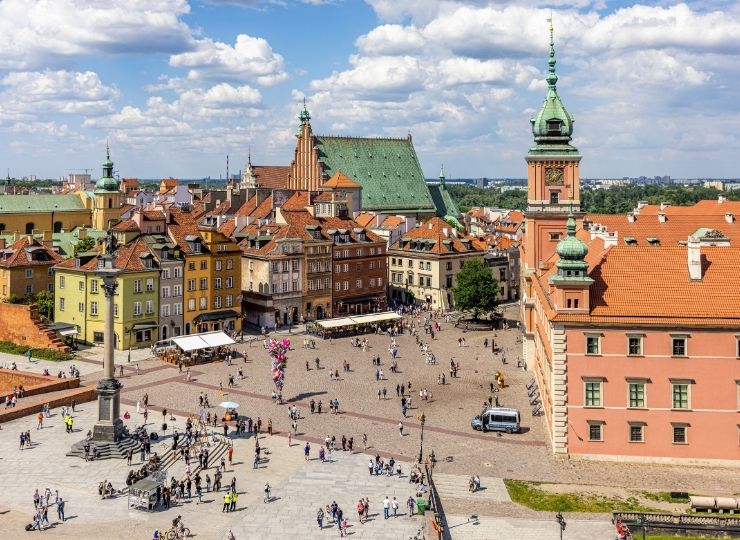A view of Castle Square in Warsaw with colorful townhouses, Sigismund's Column and the Royal Castle on a sunny day, with numerous people walking around.