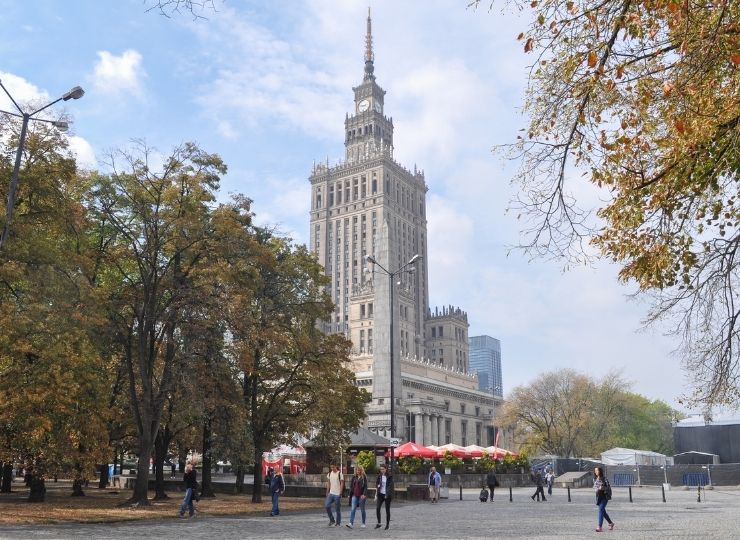 The Palace of Culture and Science in Warsaw surrounded by trees, with people walking in the park in the foreground.