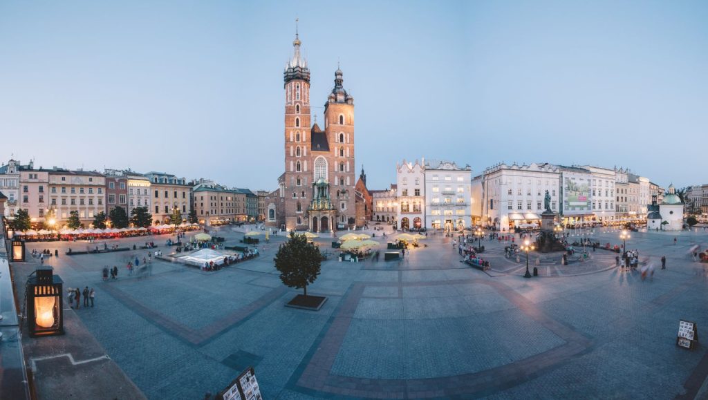 Panorama of the market square in Krakow with St. Mary's Basilica in the foreground surrounded by historic townhouses, an illuminated square and strolling people.