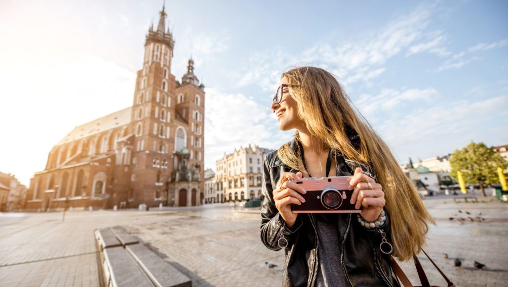 A woman with a camera in her hand smiles, standing in front of St. Mary's Basilica in Krakow.