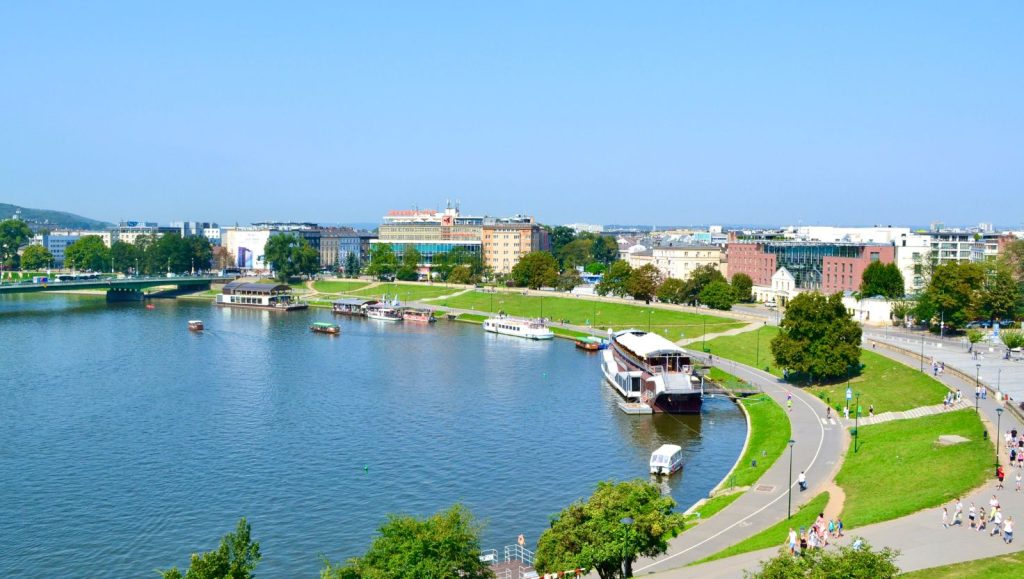 A panorama of a river with urban buildings in the background, boats and ships moored at the shore on the river, surrounded by green areas and people walking on the paths.