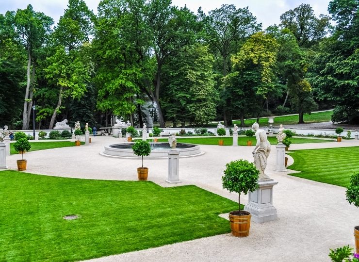 A well-maintained garden with symmetrical grass beds, marble statues, a fountain in the center and a row of trees in the background.