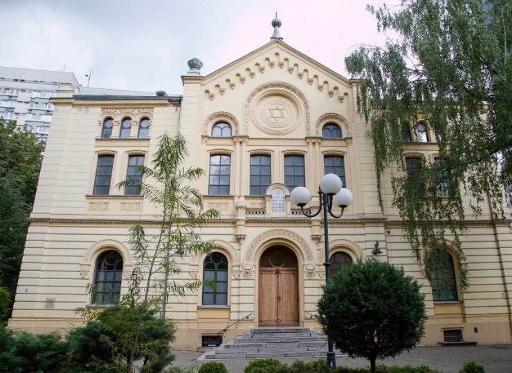 Zabytkowy budynek synagogi z kremową fasadą, dużymi oknami i drewnianymi drzwiami, otoczony zielenią, z symbolami żydowskimi na przedniej ścianie.