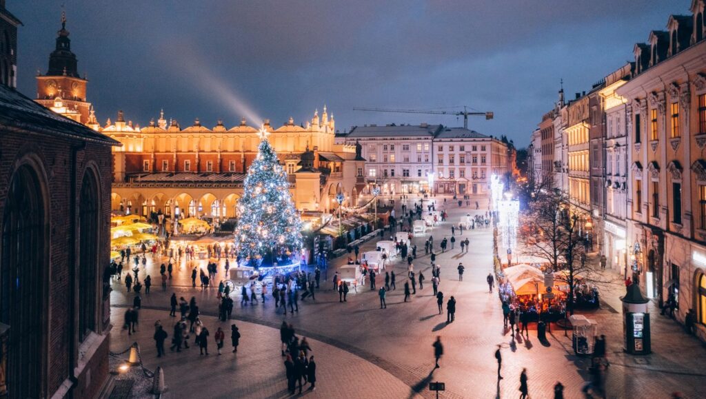 Festively illuminated Krakow Main Square with a Christmas tree in the center, surrounded by historic buildings and crowds of strolling people.
