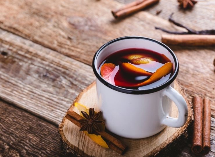 Mug of mulled wine with orange and spices on a wooden coaster, surrounded by cinnamon and star anise.