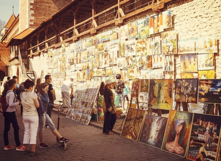A group of people views paintings displayed for sale on an exterior wall in Krakow on a sunny day.