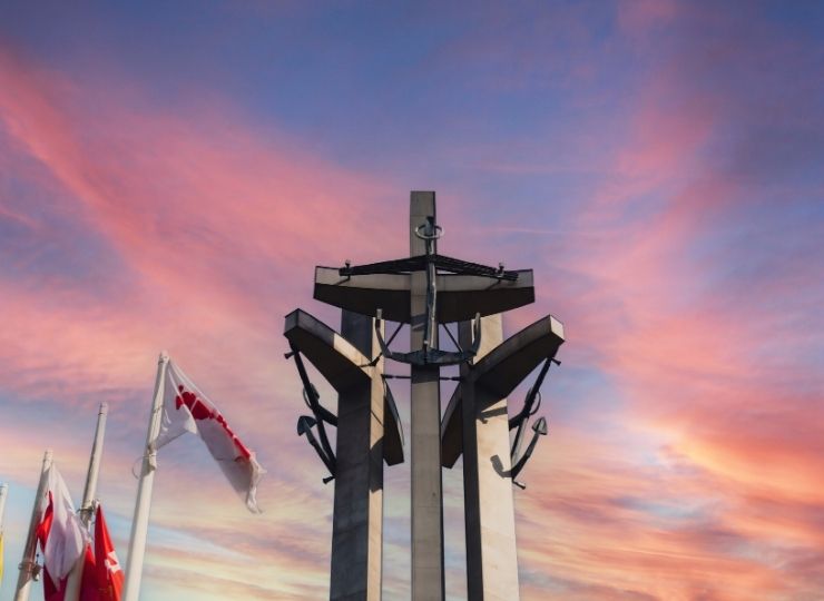 Pomnik składający się z trzech wysokich stalowych krzyży na tle kolorowego zachodu słońca, z flagami po lewej.