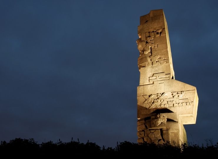 Podświetlony pomnik na Westerplatte z kamiennymi reliefami na tle ciemnego, pochmurnego nieba.