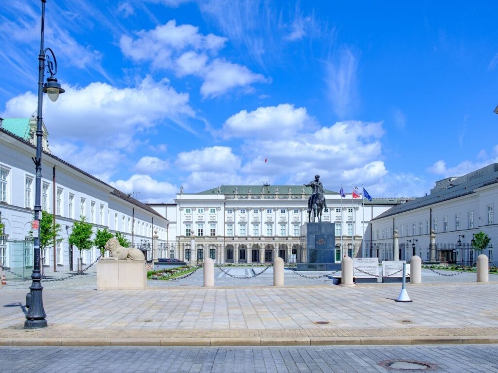 A grand neoclassical building in Warsaw,, features a central statue of a rider on horseback under a bright blue sky, flanked by symmetrical wings and stone lion sculptures.