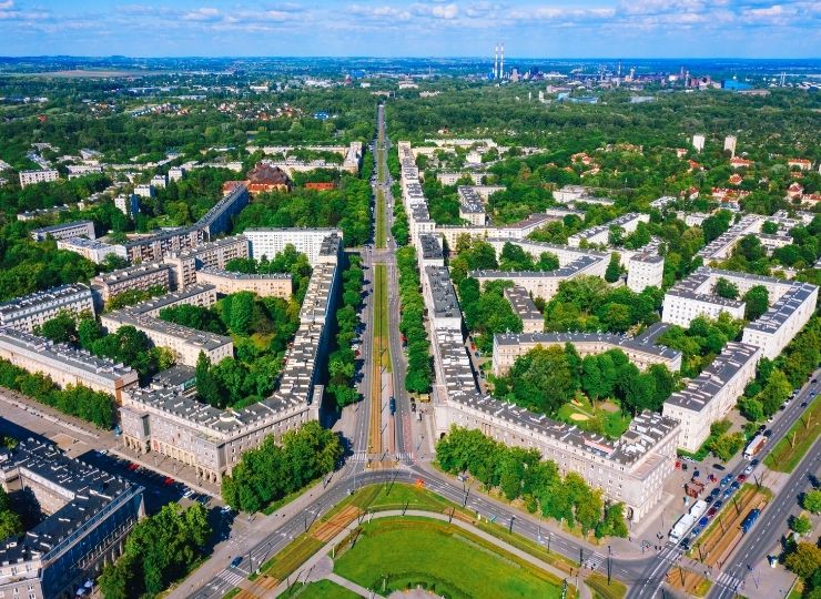 Aerial view of a city with a grid street layout, numerous residential buildings, and green spaces in the center. In the background, forests and industrial facilities are visible on the horizon.