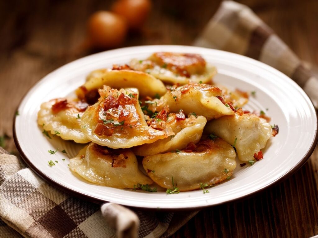 A plate of golden-brown pierogies garnished with herbs and bits of sautéed onion sits on a checkered cloth, evoking the flavors you might savor while exploring Warsaw in a day. Two blurred onions appear in the background.