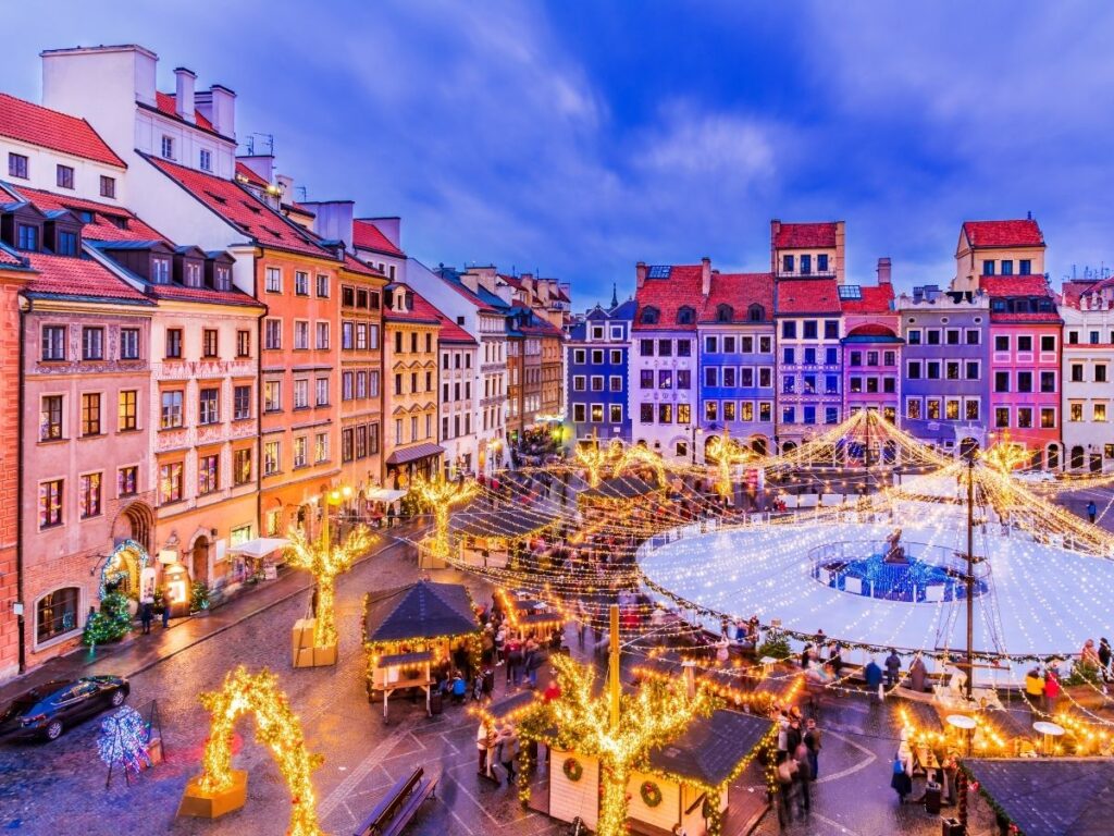 A vibrant Warsaw's old town square at dusk with colorful historic buildings, festive string lights, decorated trees, and a large circular ice skating rink surrounded by people and market stalls.