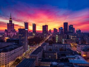 Warsaw skyline at sunset, with colorful clouds in shades of pink, orange, and blue. Tall buildings line a wide avenue as city lights begin to glow.
