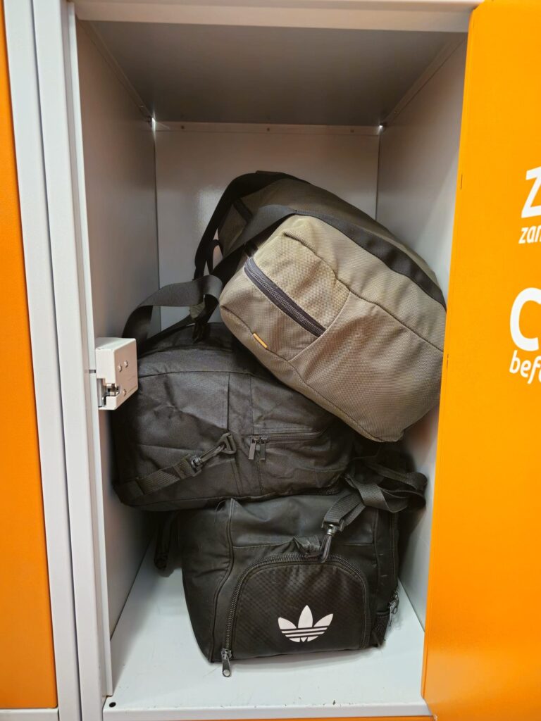 An open locker with three bags inside—depicting luggage storage in Krakow. A black Adidas duffel bag sits at the bottom, a black backpack rests in the middle, and a gray bag is stacked on top. The locker door features a bold orange color.
