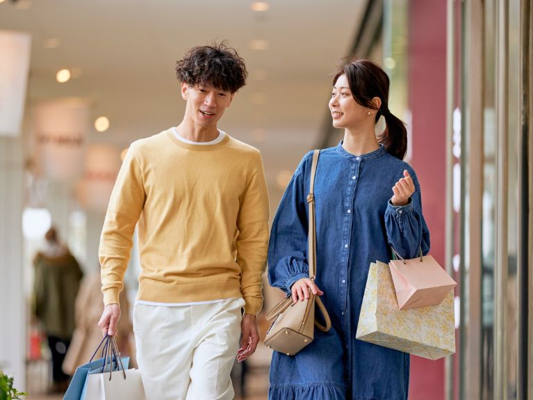 A man and a woman enter the room, smiling and carrying several shopping bags. The man is wearing a yellow sweater and white pants, while the woman is wearing a blue dress and carrying a beige handbag over her shoulder.