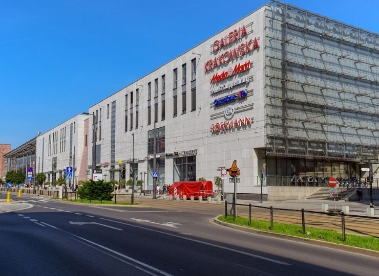 Modern Galeria Krakowska shopping mall with large store signs on the facade, including Media Markt and Rossmann, pictured next to a quiet street under a clear blue sky.
