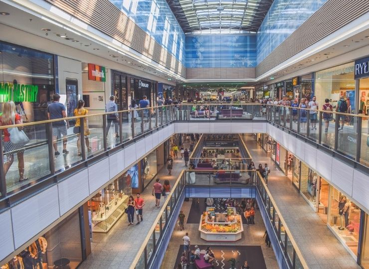 A vibrant, modern shopping mall with two floors filled with stores and people walking, shopping, and standing around. The ceiling is made of glass, letting in natural light, and numerous escalators connect the floors.