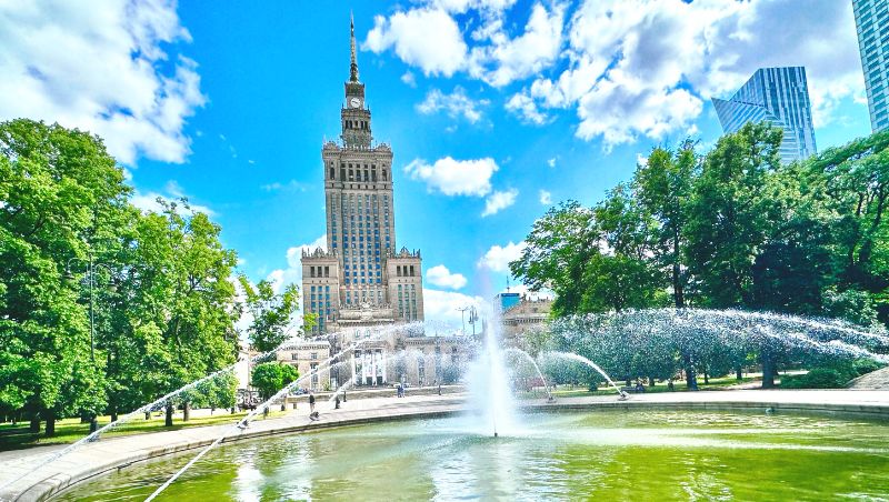 A large fountain sprays water in a circular park in Warsaw. The fountain is surrounded by trees, with a tall historic building (Palace of Culture) and modern skyscrapers in the background under a bright blue sky with scattered clouds.