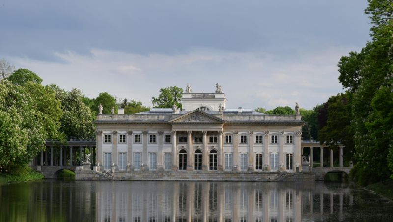 A neoclassical palace with columns and statues stands beside a tranquil lake that reflects its facade; surrounded by green trees under a partly cloudy sky.