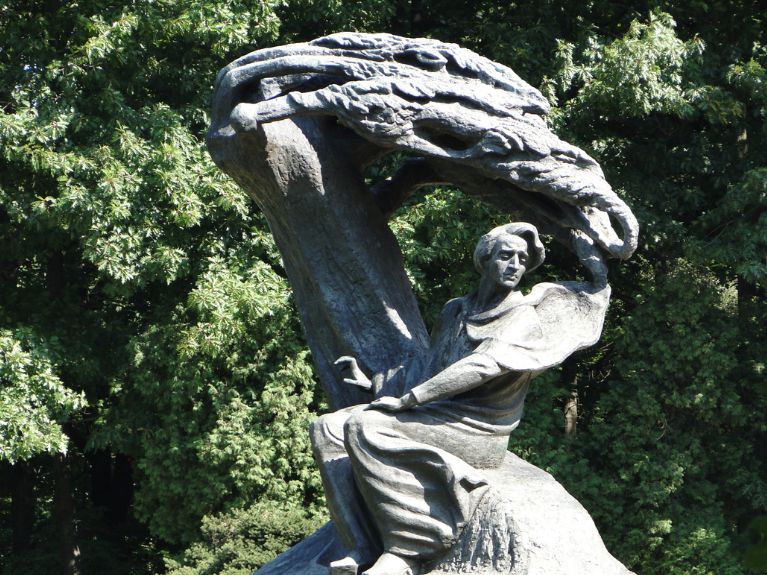 A bronze statue of Frederic Chopin as seated man in flowing robes under a dramatic arched tree, set outdoors with dense green foliage in the background.