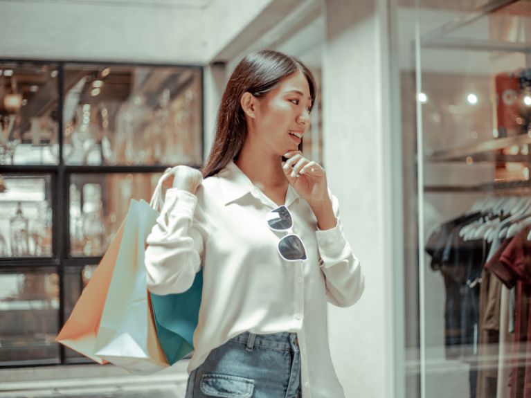 A woman with colorful shopping bags smiles while standing in front of a clothing store window, dressed in a white shirt, blue jeans, and sunglasses clipped to her shirt.