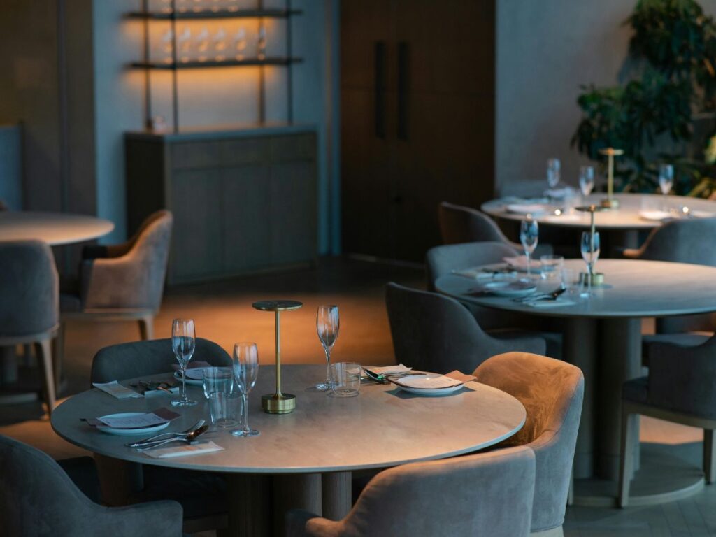 A modern, elegantly set restaurant dining room with round tables, grey upholstered chairs, wine glasses, plates, and soft lighting. Shelves with glassware and some greenery are visible in the background.