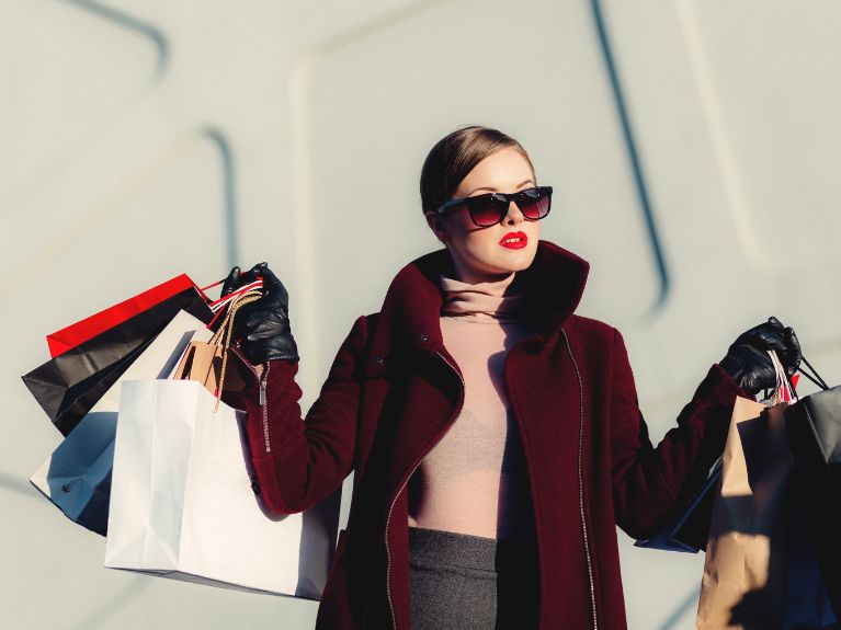 A stylish woman in sunglasses, red lipstick, a burgundy coat and black gloves holds several shopping bags in each hand against a light, textured background.