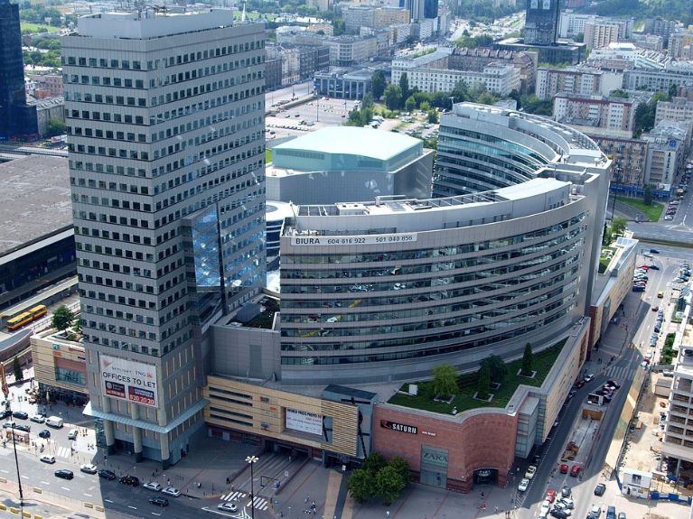 Aerial photograph of a modern cityscape with a tall rectangular skyscraper and a curved glass office building surrounded by streets, cars, and other urban structures. Greenery on the roof and several commercial signs are visible.