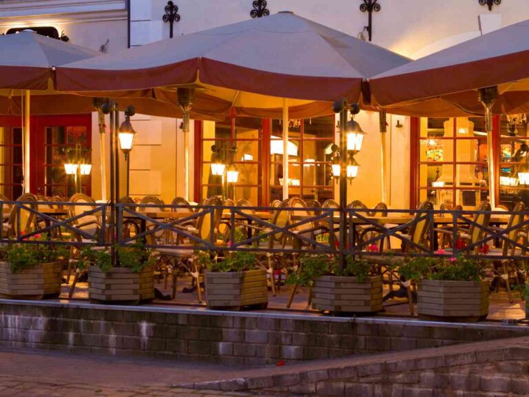 Outdoor restaurant patio in the evening, with tables and chairs under large parasols, warm lights glowing, and potted plants along the railing in front of a building with large windows.