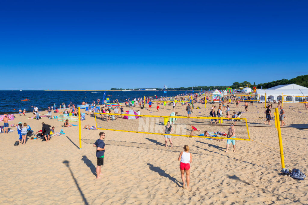 Ludzie grają w siatkówkę plażową na piaszczystej, zatłoczonej plaży pod bezchmurnym, błękitnym niebem. Wielu innych odpoczywa, opala się i pływa. W tle, przy linii brzegowej, rozstawione są namioty i kolorowe parasole.