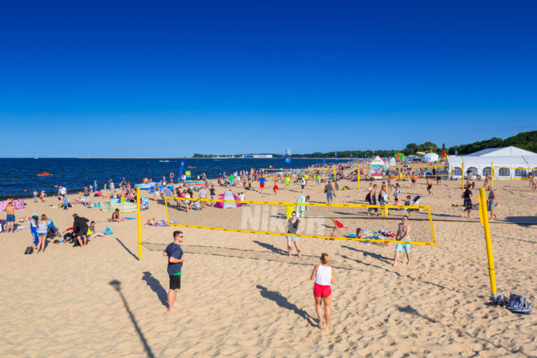 Ludzie grają w siatkówkę plażową na piaszczystej, zatłoczonej plaży pod bezchmurnym, błękitnym niebem. Wielu innych odpoczywa, opala się i pływa. W tle, przy linii brzegowej, rozstawione są namioty i kolorowe parasole.