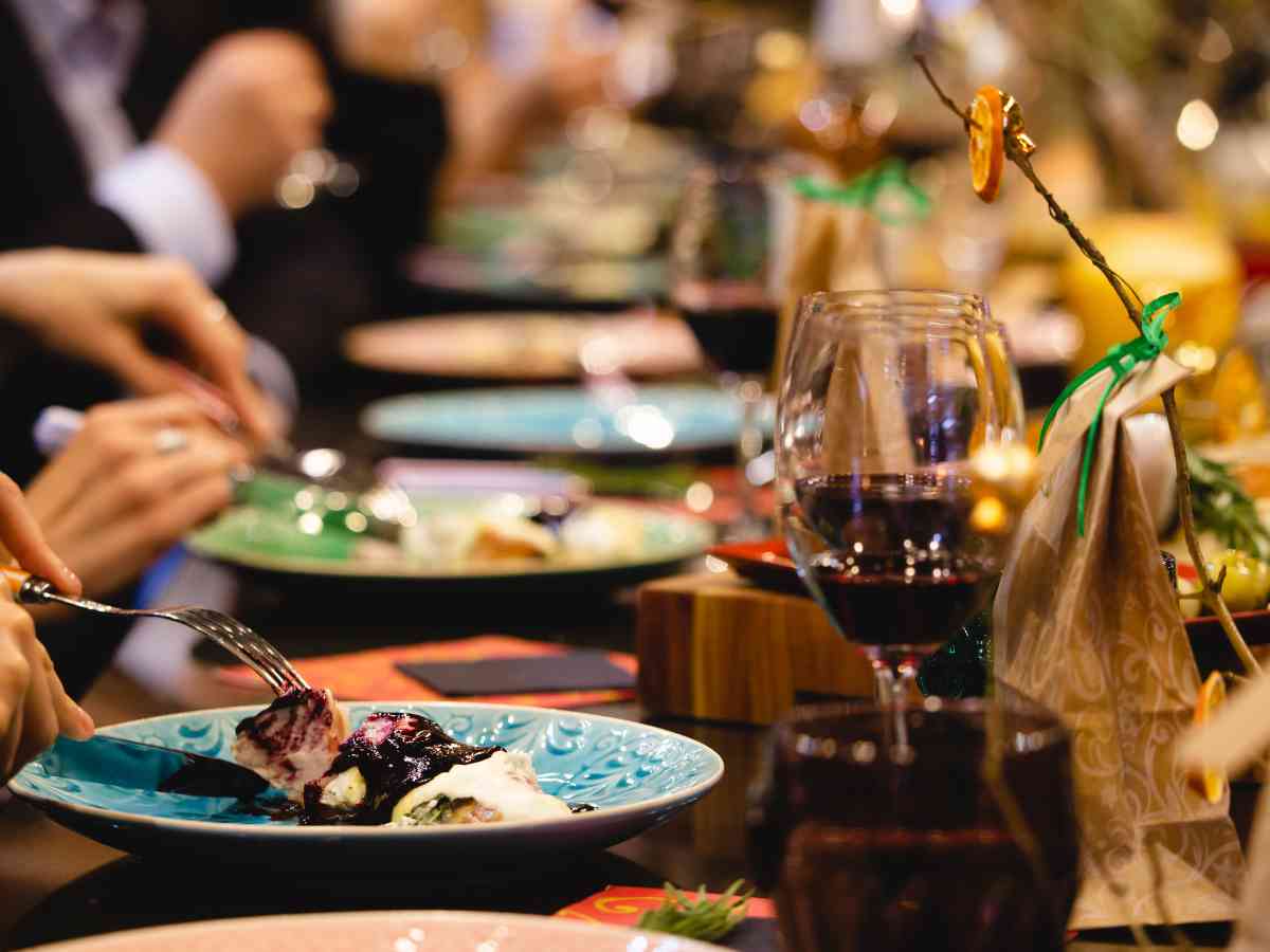 A close-up of a festive dining table with plates of food, glasses of red wine, and people’s hands using cutlery, suggesting a social meal or celebration.