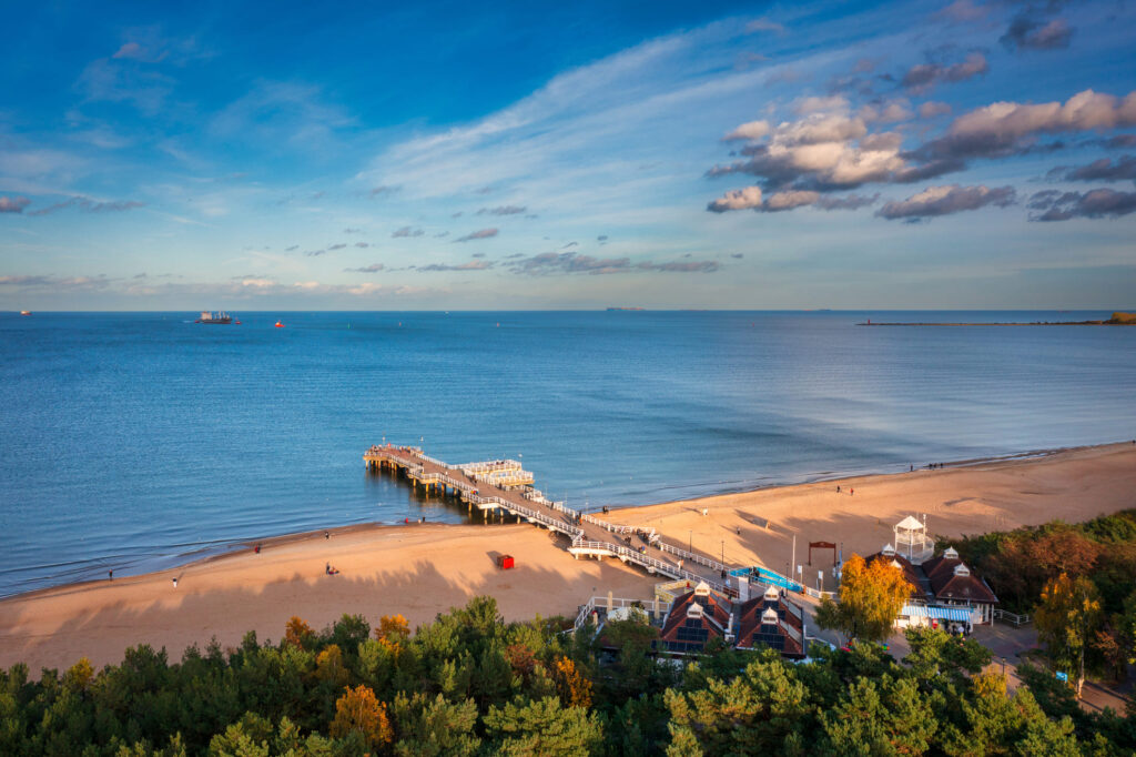 Piaszczysta plaża z drewnianym pomostem wychodzącym w spokojne, błękitne wody, otoczona zielonymi drzewami i małymi budynkami pod częściowo zachmurzonym niebem. Ludzie spacerują po pomoście i plaży, a na horyzoncie widoczne są statki.