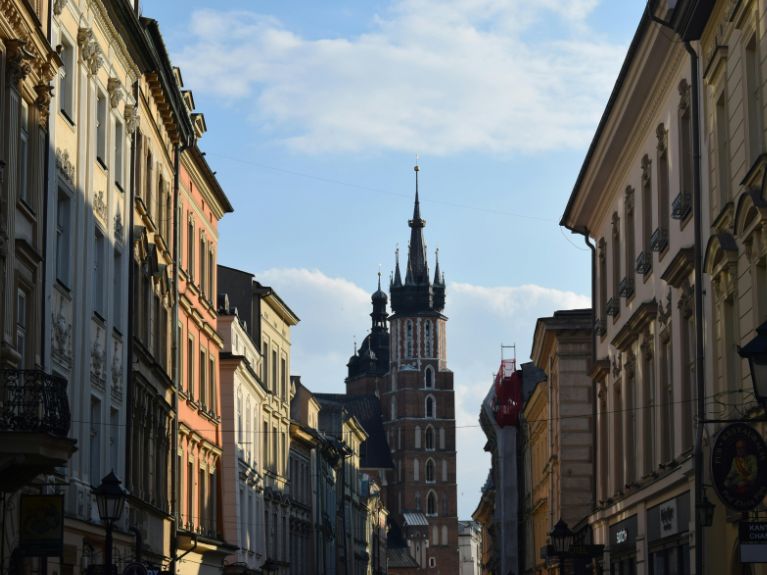 A tall brick church with twin spires rises above a narrow street lined with historic buildings in Cracow under a partly cloudy blue sky.